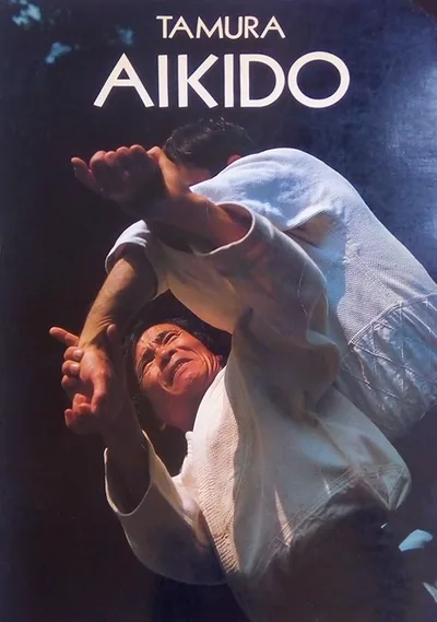 Couverture d’Aïkido (1986) : « TAMURA » et « AIKIDO » en capitales blanches au-dessus d’une photo en couleur d’une projection, deux aïkidokas en keikogi blanc sur fond sombre