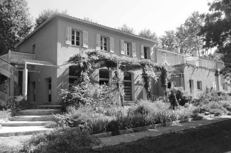 Photo en noir et blanc du Shumeikan à Bras : une bâtisse provençale à un étage avec des volets, une pergola couverte de vigne le long du rez-de-chaussée, des marches en pierre et un jardin planté devant, de grands arbres derrière.